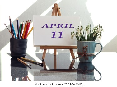 Calendar for the eleventh of April: an easel with the inscription April in English and the number 11, a bouquet of snowdrops in a cup, multi-colored pencils, brushes in the sun from the window