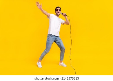 Foto de cuerpo entero de un buen chico brunet milenario que canta, usa gafas, camiseta, jeans, zapatillas deportivas aisladas en fondo amarillo