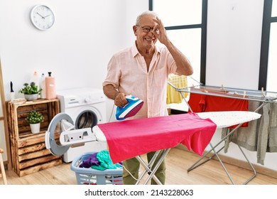 Senior man ironing clothes at home smiling happy doing ok sign with hand on eye looking through fingers 