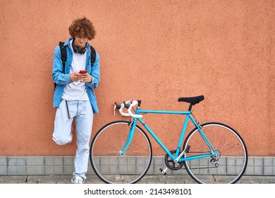 Jonge curly-haired student met koptelefoon staande op de muur buiten met behulp van mobiele telefoon naast haar blauwe fiets
