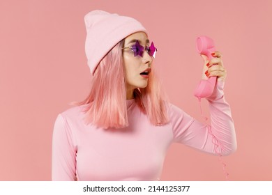 Joven mujer sorprendida con el pelo de rosa teñido brillante en gafas de sombrero de camisa rosa sostiene un teléfono vintage aislado en un retrato de estudio de fondo rosa pastel claro. Concepto de moda de estilo de vida de personas