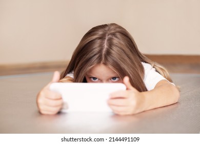 Kleines Mädchen benutzt Smartphone, schaut auf den Bildschirm, spielt oder schaut Zeichentrickfilme und liegt auf dem Boden. Kind mit Gadget, Modell