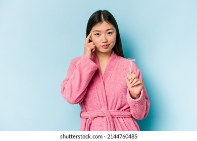 Wanita muda asia memegang pisau cukur terisolasi di latar belakang biru menunjuk kuil dengan jari, berpikir, fokus pada tugas.