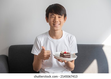 Surprised and happy asian teen boy with  chocolate cake in a plate sit on sofa in room.