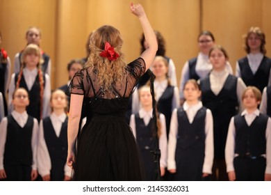 Vrouw zanger blonde in zwarte jurk met opgeheven hand leidt het koor staande voor een groep kinderen studenten van een muziekschool achteraanzicht van de leider