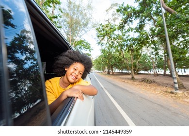 Little African child girl sitting in the car with pull her face and hand out of the window in summer sunny day. Happy Asian family enjoy and fun together with outdoor lifestyle on road trip vacation. 