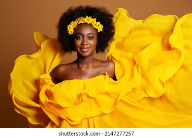 Glad mørkhudet kvinde i gul modekjole. Skønhed afroamerikansk model danser i silkekjole vinker Flyver på vinden over beige baggrund. Gul blomsterkrans i kvinder sort krøllet frisure