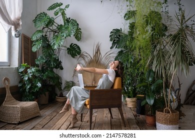 Mulher freelancer sobrecarregada sentada em uma cadeira em uma estufa aconchegante, descansando, esticando os braços com os olhos fechados. Mulher sorridente e cansada em seu jardim doméstico, fazendo uma pausa nos estudos online. Conceito para amantes de plantas.