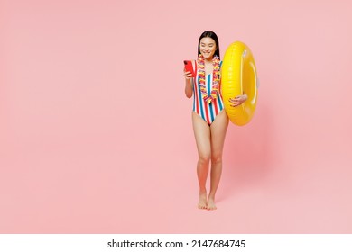 Full body young woman of Asian ethnicity in striped one-piece swimsuit lei hold inflatable ring mobile cell phone isolated on plain pastel pink background. Summer vacation sea rest sun tan concept.