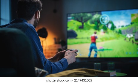 Un joven pasa el tiempo en casa, sentado en un sofá en un elegante apartamento tipo loft, jugando a un videojuego arcade de disparos en una consola. Un hombre usa un mando para jugar a un juego multijugador masivo en línea de estilo Battle Royale.