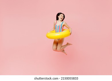 Jeune femme d'origine asiatique en maillot de bain une pièce rayé avec anneau gonflable jaune et collier hawaïen sautant haut dans l'eau isolé sur fond rose pastel uni. Concept de repos en mer pour les vacances d'été