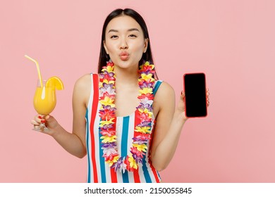 Junge Frau asiatischer Abstammung in gestreiftem einteiligem Badeanzug Hawaii Lei hält Cocktail-Handy mit leerem Bildschirm isoliert auf schlichtem pastellrosa Hintergrund. Sommerurlaub Meer Ruhe Sonnenbräune Konzept