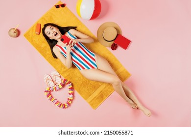 Draufsicht Ganzkörper-junge Frau asiatischer Abstammung in gestreiftem Badeanzug liegt auf Handtuch-Hotel-Pool-Haltegriff, Handy isoliert auf einfarbigem rosa Hintergrund. Sommerurlaub Meer Ruhe Sonnenbräune Konzept