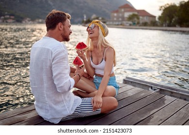 Ein Paar hat eine gute Zeit bei einem Date am Meer, das auf einem Holzsteg sitzt, lacht und Wassermelone isst. Liebe, Spaß, Zusammengehörigkeitskonzept.