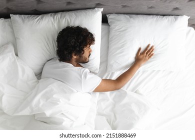 Top view of lonely young indian guy laying in bed alone, touching empty pillow next to him, experiencing difficult break up or divorce, feeling lonely, bedroom interior, copy space