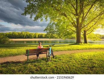 Frau sitzt auf Bank und Mountainbike, grüne Bäume und See bei Sonnenuntergang im Frühjahr. Bunte Landschaft mit stillstehendem Mädchen, Fahrrad, Fluss, grünem Gras, Fluss im Park. Sommer. Sport und Reisen. Radfahren