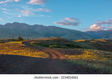 Scenic view from sunlit grassy hill with dirt road to high mountain range. Beautiful sunny mountain landscape with large mountains. Colorful scenery with hill in sunlight against awesome mountains.