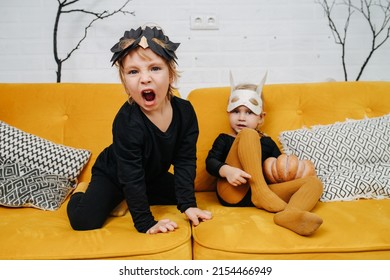 Playful awkward preschooler kids dressed for halloween on a yellow couch, wearing masks. Boy is roaring like a lion. Room decorated with dead leafless branches.