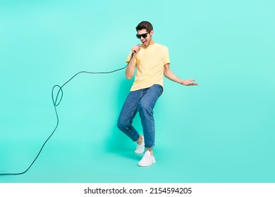 Foto de cuerpo entero de un joven brunet divertido que canta con gafas, pantalones vaqueros, calzado aislado en un fondo turquesa