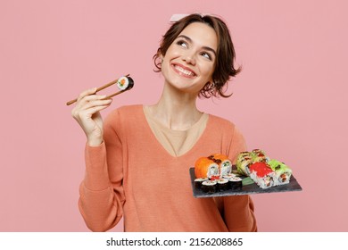 Young minded fun woman in sweater hold makizushi sushi roll served on black plate traditional japanese food eat roll with chopsticks look aside on workspace isolated on plain pastel pink background.