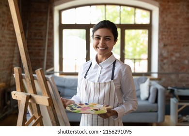 Drăguță pictor indiană pozează în atelier lângă șevalet cu pânză ține paleta și zâmbet cu pensula Privește camera, bucură-te de studiu în școala de artă și hobby creativ. Concept de portret de artist profesionist