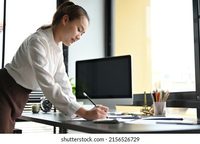 Erfolgreiche asiatische Geschäftsfrau, die sich an ihren Schreibtisch lehnt und etwas auf die Berichtsunterlagen schreibt.