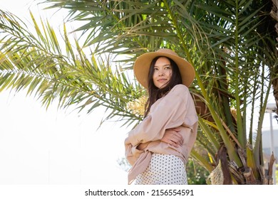 ภาพมุมต่ำของหญิงสาวผมสีน้ำตาลเข้มที่ดูมีความสุข ยืนกอดอกอยู่ใกล้ต้นปาล์ม