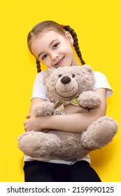 Cô gái nhỏ dễ thương ôm một con gấu bông khi ngồi trên nền màu vàng. Một cô bé xinh đẹp với bím tóc, nụ cười và vẻ ngoài ngoan hiền. Cô gái mỉm cười hạnh phúc với đồ chơi gấu bông