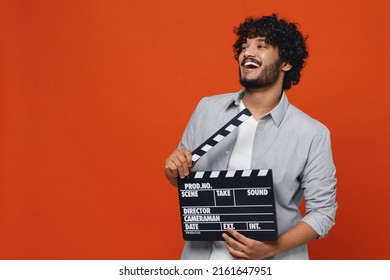 Presretan, uzbuđen, smije se likujući, zadivljujući mladi bradati Indijac, 20-godišnjak, nosi plavu košulju i drži klasičnu crnu filmsku klapu, izoliranu na običnoj narančastoj pozadini, studijski portret