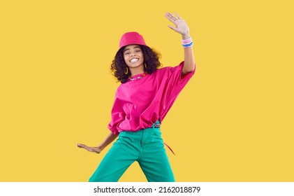 Happy joyful Black kid in funky outfit dancing in studio. Cheerful African dancer girl wearing loose fuchsia top, bucket hat and green pants enjoying hype and having fun. Children's fashion concept