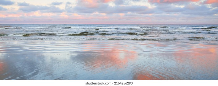 Pemandangan panorama laut Baltik dari pantai berpasir. Langit matahari terbenam yang cerah, awan merah muda bercahaya berwarna-warni, cahaya lembut. Refleksi simetri pada air, cermin alami. Cuaca, musim dingin, perubahan iklim, alam