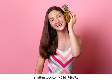 Fröhliches, lächelndes, gebräuntes Teenager-Mädchen mit langen, glänzenden, dunkelbraunen Haaren, das eine Ananas wie ein Handy hält, trägt einen bunt gestreiften, einteiligen Badeanzug, der auf rosafarbenem Hintergrund isoliert ist, Haarpflegekonzept