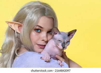 頬の近くにスフィンクス猫を保持しているコスプレ エルフの若い女性の肖像画。夏の青いドレスを着たエルフ、美しい金髪の巻き毛の長い髪、さまざまな色の目、ピアスの耳。黄色の背景。