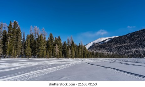 Jejak ban yang berpotongan terlihat di permukaan danau beku yang tertutup salju. Hutan jenis konifera di pantai. Sebuah gunung melawan langit biru. Altai. Danau Multinskoe Bawah