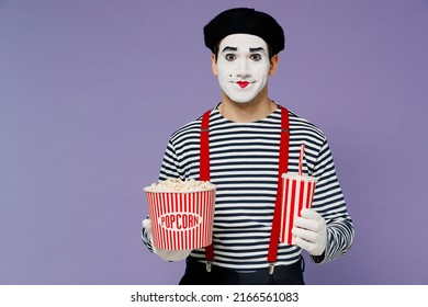 Charismatischer, erstaunlicher, wunderbar lebhafter junger Mime-Mann mit weißer Gesichtsmaske trägt gestreiftes Hemd, Baskenmütze, alten Eimer Popcorn, Tasse Limonade, isoliert auf schlichtem pastellfarbenem hellviolettem Hintergrund, Studioporträt