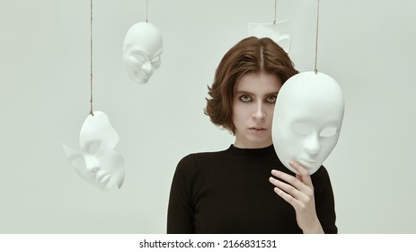 Teatro de la vida. Una chica vestida de negro se encuentra en una habitación blanca rodeada de varias máscaras mirando fijamente a la cámara. Hipocresía. Roles humanos. Desordenes mentales.