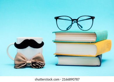 White mug with a felt moustache and silcy bow tie on the blue background with stack of books and glasses. Copy space. Concept of Father's day.