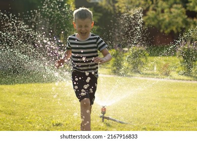 A little wet boy runs barefoot on the lawn next to the sprinkler. Happy carefree childhood and holidays concept. 