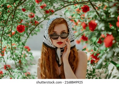 Hermosa mujer pecosa de pelirroja de moda con gafas de sol de ojos de gato de estilo retro de moda, pañuelo en la cabeza con estampado de lunares, posando en un jardín de rosas rojas. Copiar, espacio vacío para texto