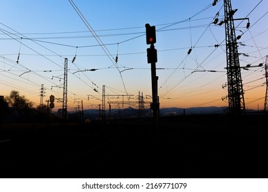 夕暮れの暗い背景に鉄道の赤信号セマフォ。夕焼け空を背景に黒いシルエットのカテナリーとマスト
