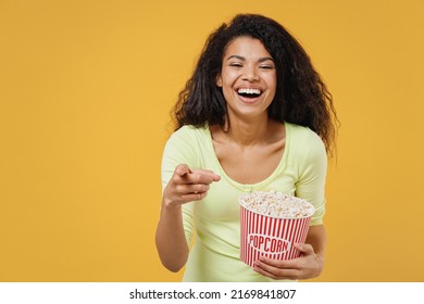 Schöne, lächelnde, lachende, faszinierende afroamerikanische junge Frau in den Zwanzigern trägt grünes Hemd, sieht sich einen Film an, hält einen Eimer Popcorn, zeigt mit dem Finger auf die Kamera, isoliert auf gelbem Hintergrund, Studioporträt.