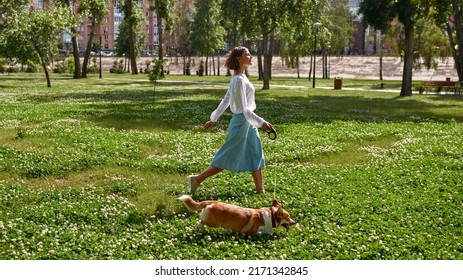 Vista laterale di una giovane donna caucasica sorridente che porta a spasso un grazioso cane Corgi su un prato verde nel parco. Concetto di relazione tra uomo e animale. Idea di amicizia tra proprietario e animale domestico. Giornata estiva soleggiata