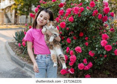 Linda niña de 6 a 7 años sosteniendo un gato posando sobre florecientes arbustos de rosas en la calle de la ciudad al aire libre. Mirando a la cámara. Infancia. Temporada de verano. Amistad.