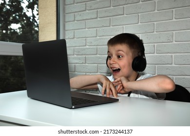 6～7 歳の男の子が窓際の白いテーブルに座り、大きなヘッドホンをつけてノートパソコンの画面をじっと見つめている