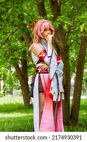 Porträt eines schönen Teenager-Mädchens im YAEMIKO-Anime-Kostüm im Park im Freien. Figur der Computerspielgöttin mit rosa Haaren YaeMiko. Mädchen, das als Anime-Held verkleidet ist, steht in der Nähe von Baumsommerferien
