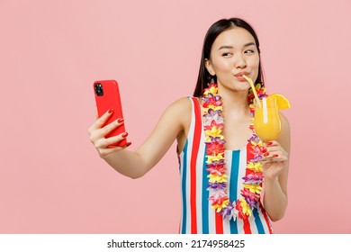 Junge Frau asiatischer Abstammung in gestreiftem einteiligem Badeanzug hawaii lei hält Cocktail und macht Selfie-Aufnahme auf Mobiltelefon isoliert auf schlichtem pastellrosa Hintergrund. Konzept der Sommerferien am Meer.