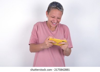 Caucasian woman with short hair wearing pink T-shirt over white wall holding in hands cell playing video games or chatting