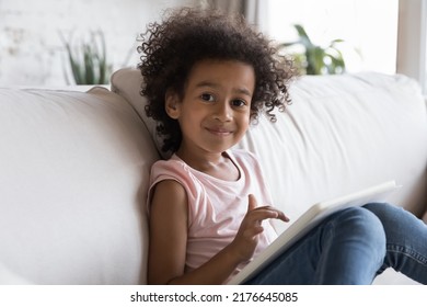 Smiling little African girl use digital tablet play new on-line game spend leisure sit on sofa at home pose for camera. Addicted to modern technology of young generation, parental control apps concept