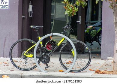 Sepeda hitam dan kuning 10 kecepatan terkunci pada struktur parkir logam melingkar. Itu di trotoar dan di depan jendela reflektif. Pengaturan perkotaan.