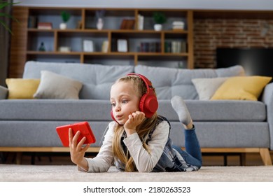 Gadis kecil yang terkejut mengenakan earphone merah tergeletak di karpet di rumah. Gadis imut memegang smartphone, menonton kartun, mengirim pesan teks, menjelajahi internet dan media sosial, bermain game